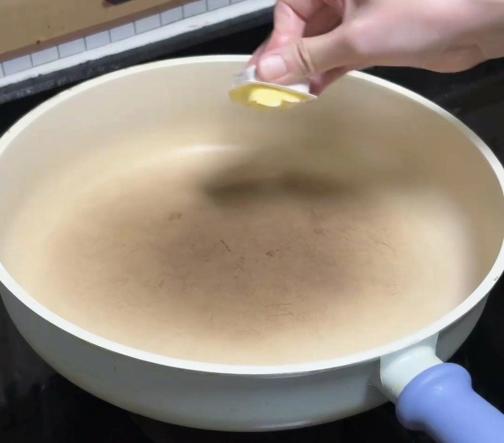 Una mano che mette un cubetto di burro giallo in una padella pulita in ceramica bianca sul fuoco.