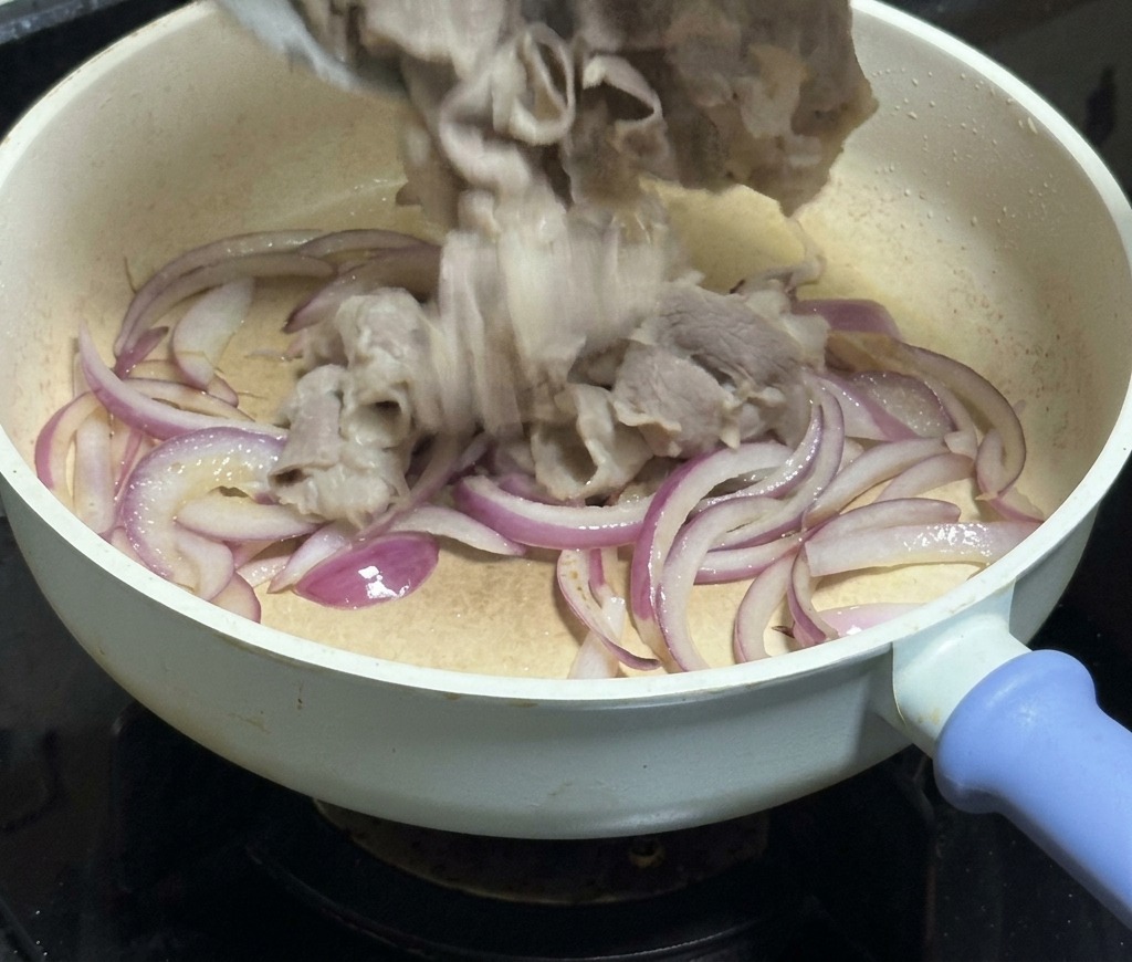Manzo a fettine sottili versato da una ciotola bianca a una padella piena di strisce di cipolla rossa traslucida.