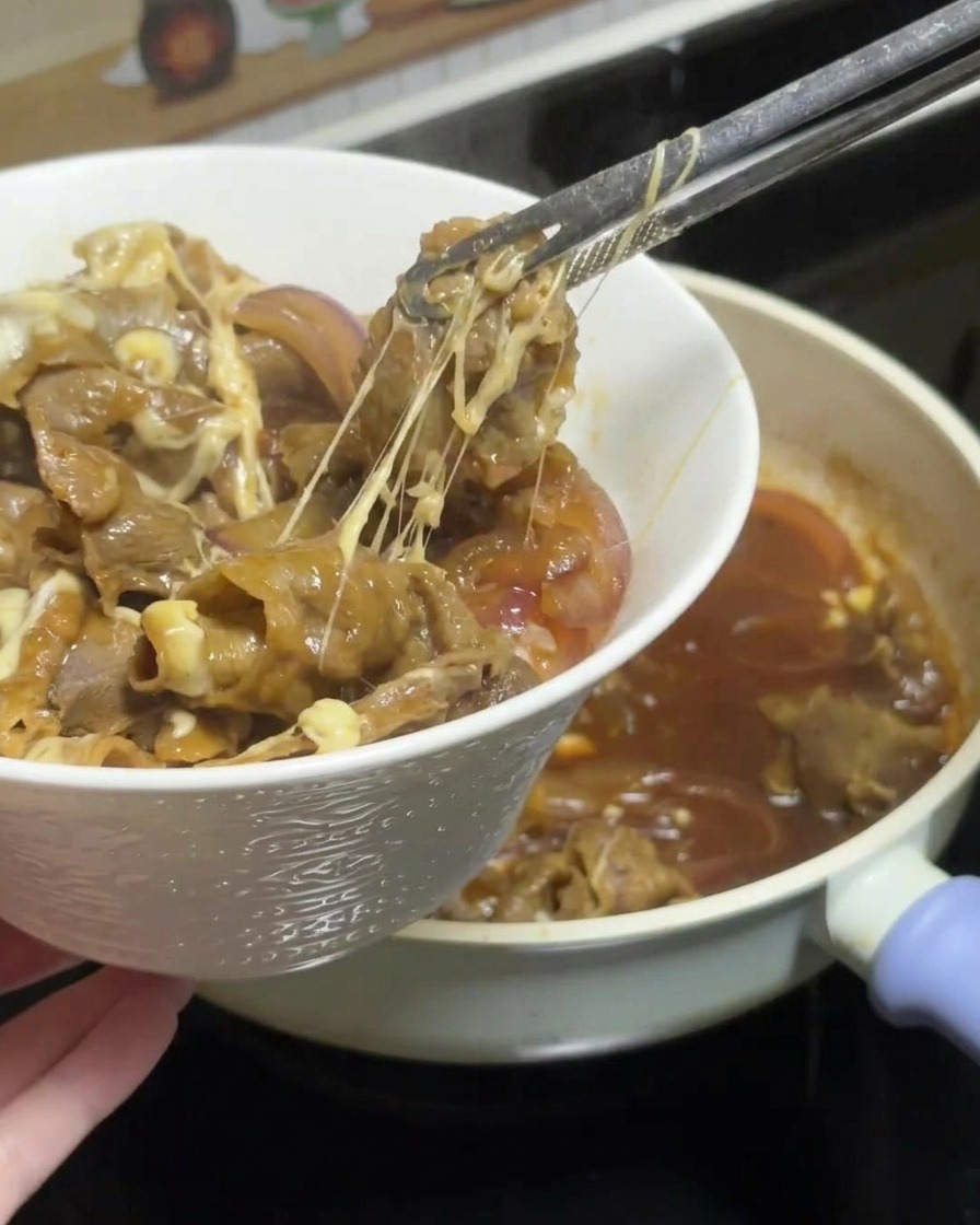 Pinze che sollevano una porzione di manzo teriyaki filante e cipolle dalla padella, mostrando lunghi fili di formaggio, e la adagiano su una ciotola di riso bianco.