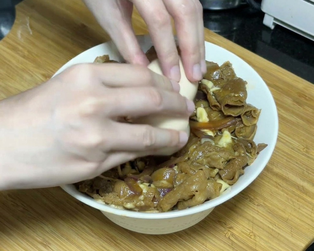 Due mani che rompono con cura un uovo onsen morbido sopra una ciotola di manzo teriyaki coperta di formaggio fuso.