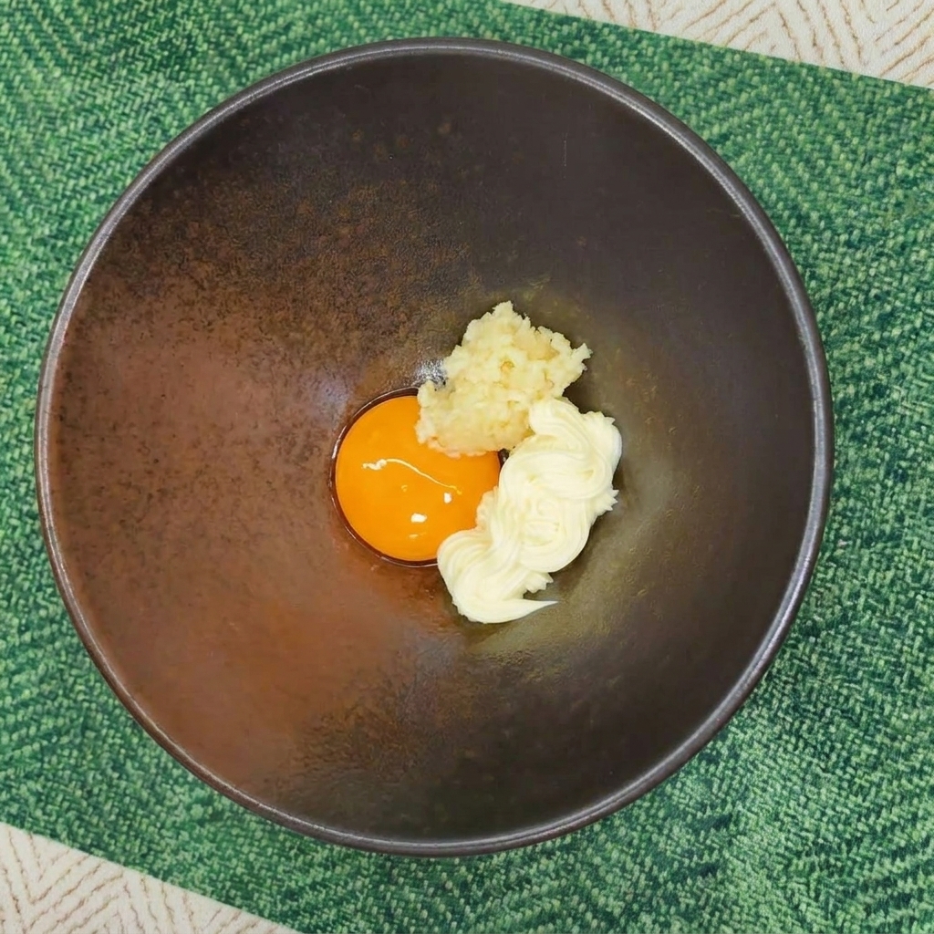Una ciotola di ceramica scura contenente un tuorlo duovo, una dose di maionese e aglio tritato su una tovaglietta verde.