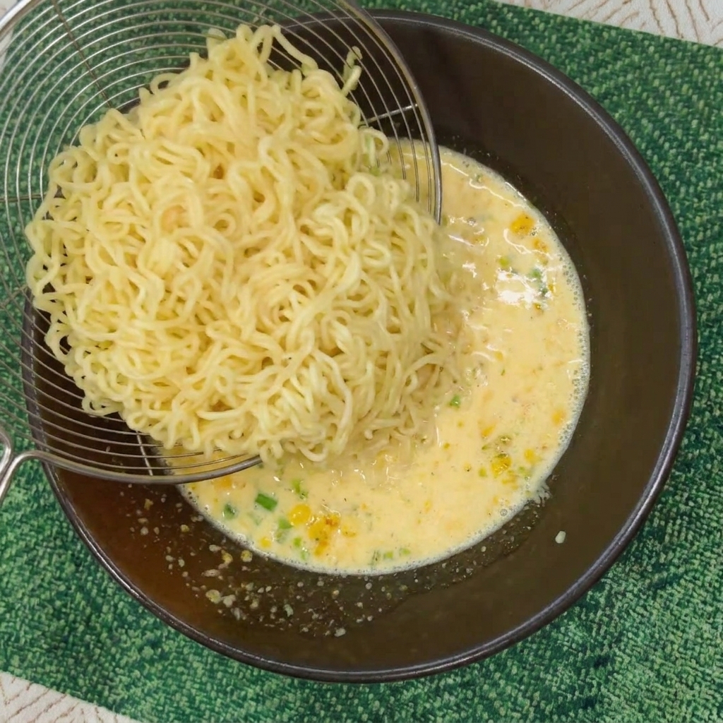 Uno scolapasta che trasferisce noodles istantanei ondulati in una ciotola colma di brodo giallo cremoso.