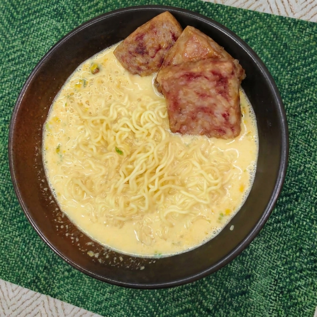 Noodles ondulati in brodo cremoso guarniti con tre fette di spam ben rosolate in una ciotola scura.