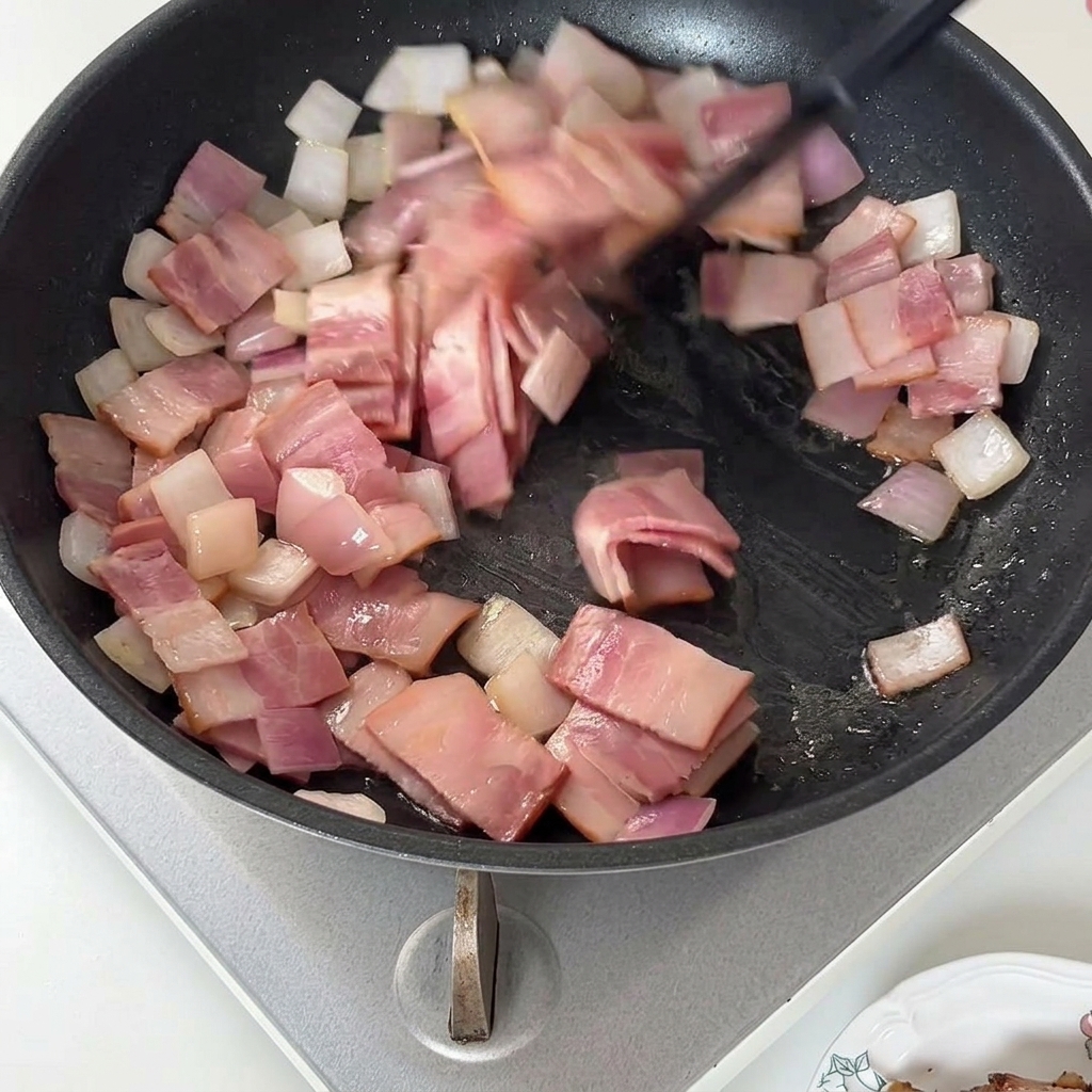 Una spatola che mescola pezzetti di bacon cotto e cipolle in una padella.