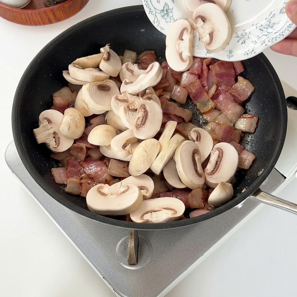 Fette di funghi champignon bianchi aggiunte in una padella calda con bacon e cipolla già cotti.