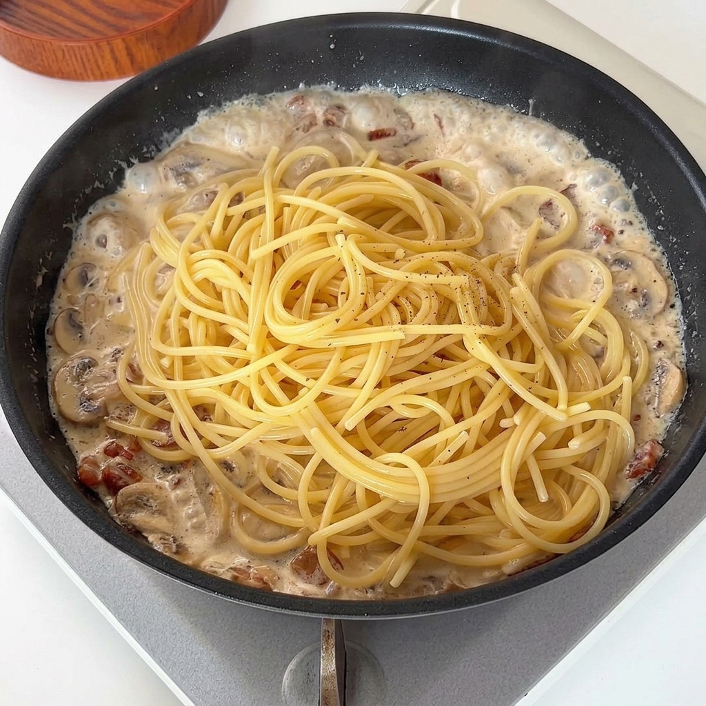 Spaghetti cotti aggiunti sopra la salsa cremosa ai funghi e bacon in padella, rifiniti con pepe nero.