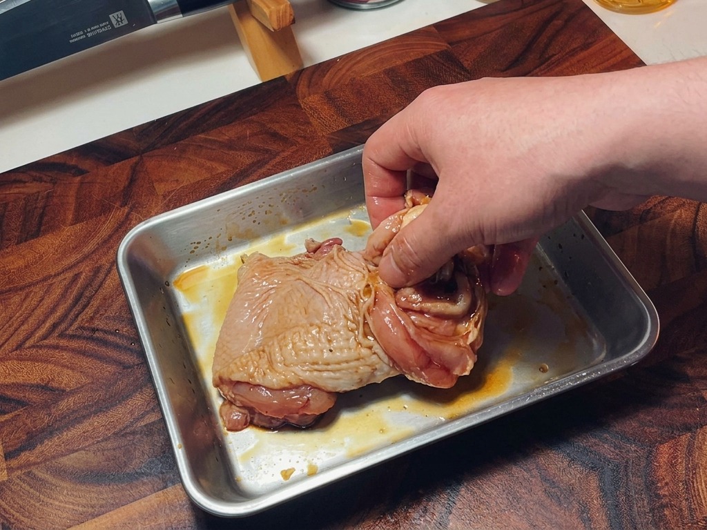 Una mano che mescola le cosce di pollo crudo condite in un vassoio per distribuire uniformemente la marinatura.