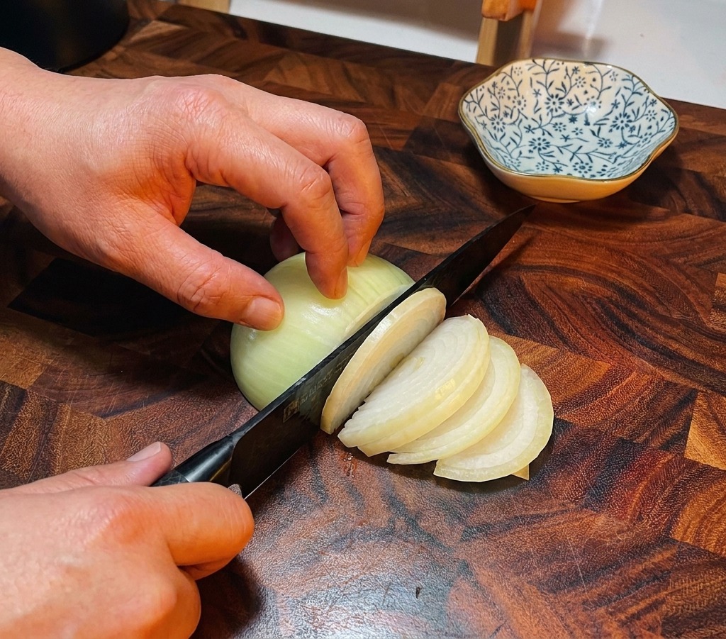 Un coltello da chef che affetta una cipolla bianca su un tagliere di legno scuro.