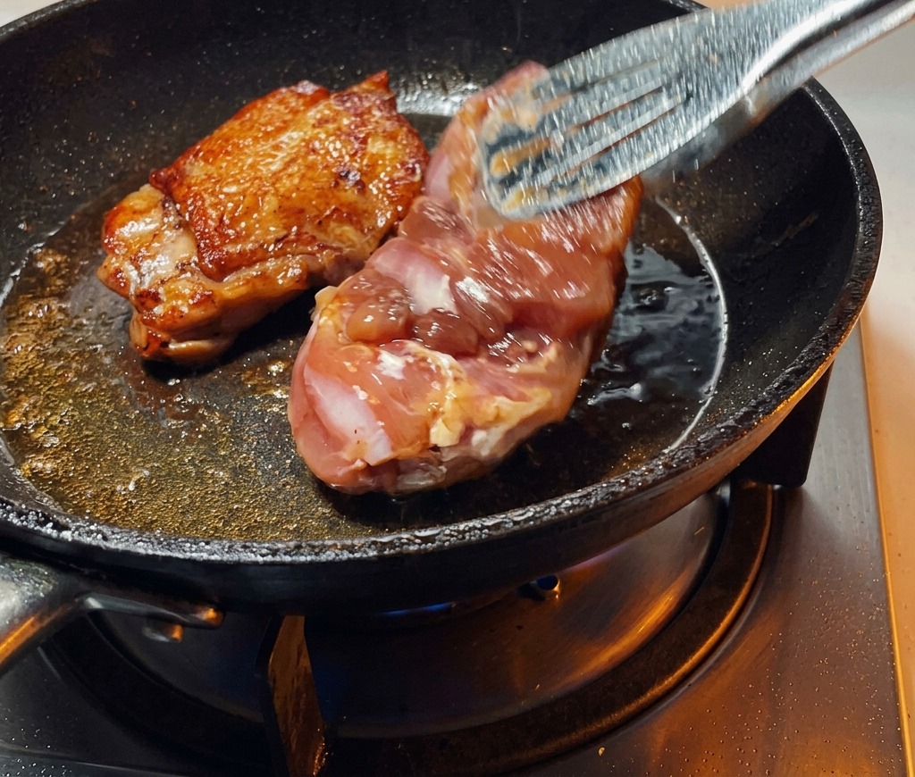 Pinze da cucina che girano una coscia di pollo scottata in padella, mostrando una pelle perfettamente dorata e croccante.