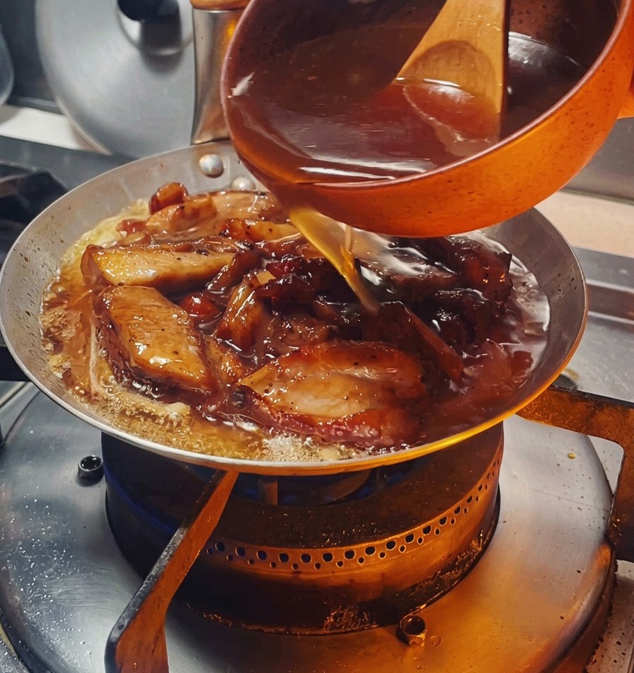 Una salsa scura e saporita versata da una piccola ciotola arancione in una padella piena di pezzi di pollo dorati e cipolle.