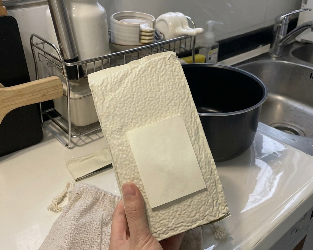 Una mano tiene un sacchetto bianco e versa il riso a chicco corto nel cestello interno nero di un cuociriso su un piano cucina pulito.