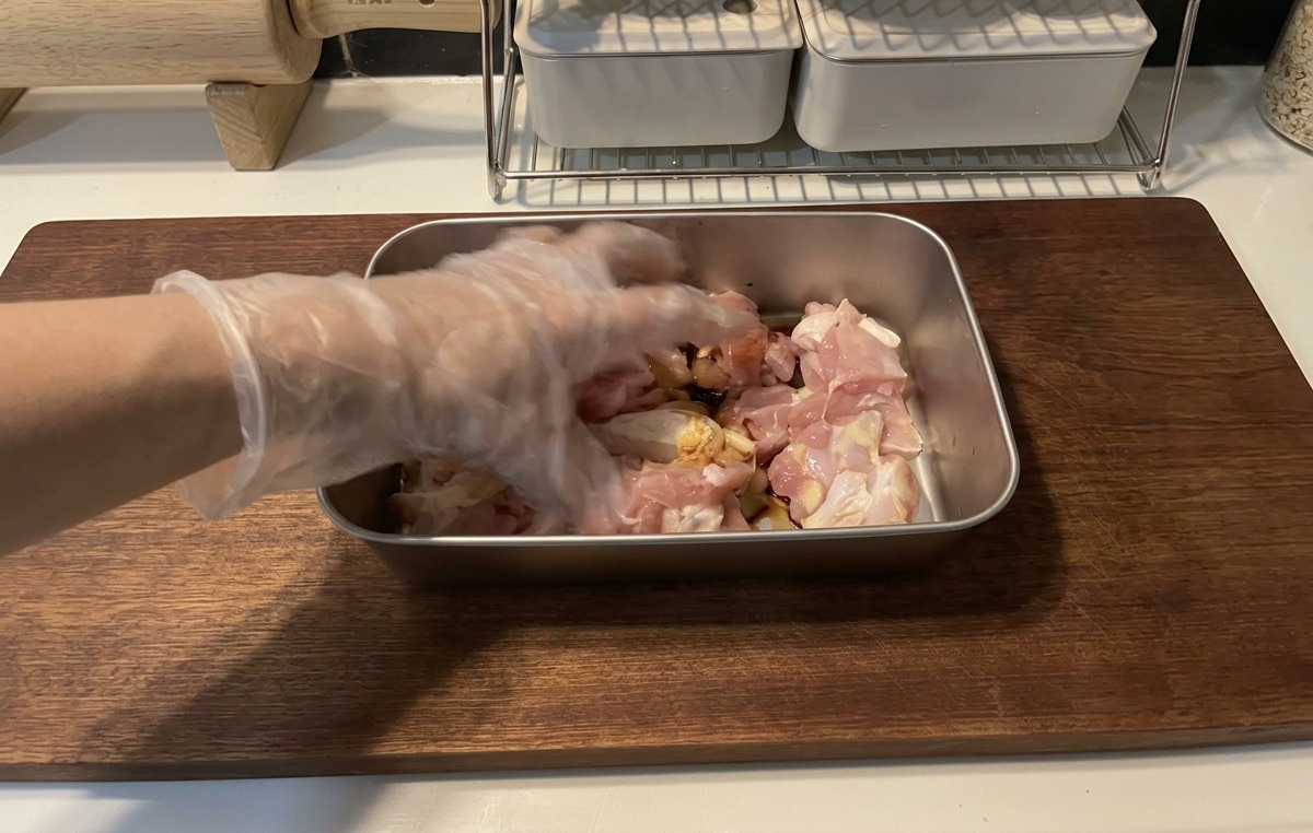 Una mano con un guanto di plastica trasparente mescola i pezzi di pollo crudo con una marinatura scura alla salsa di soia in un vassoio di metallo su un tagliere di legno.