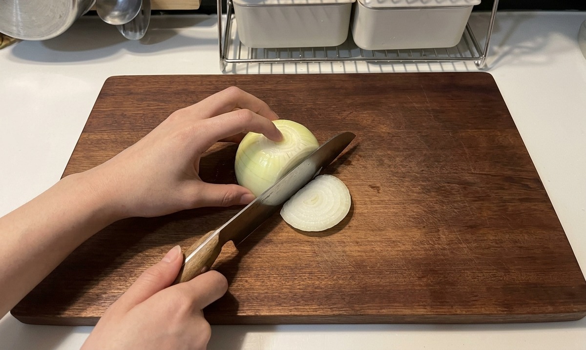 Mani che usano un coltello con manico in legno per affettare una cipolla bianca in sottili strisce a mezzaluna su un tagliere di legno scuro.