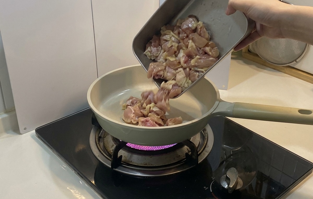 Pezzi di pollo marinato versati da un vassoio di metallo in una padella in ceramica chiara sopra la fiamma viola di un fornello a gas.