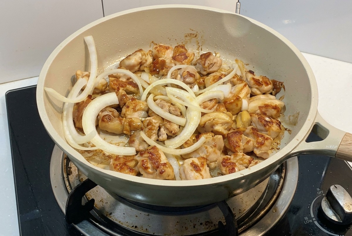Anelli di cipolla bianca affettata aggiunti a una padella con pezzi di pollo che si stanno dorando sul fuoco.