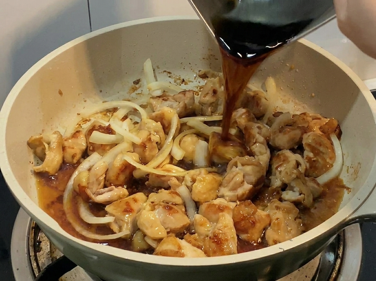 Un liquido scuro e saporito viene versato da un contenitore di metallo in una padella piena di pollo e cipolle.