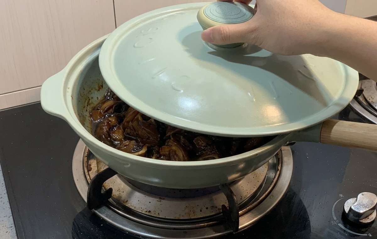 Una mano mette un coperchio azzurro su una padella verde chiaro contenente pollo e cipolle che sobbollono in una salsa scura.