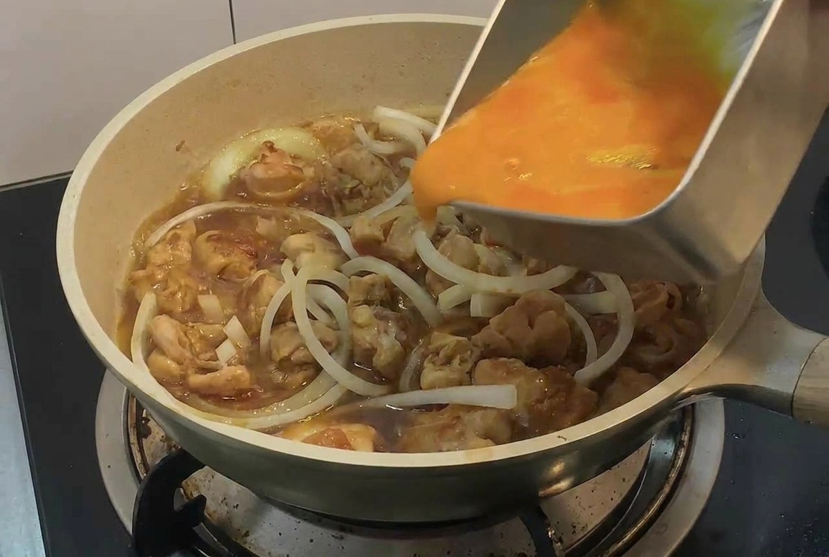 Una mano versa luovo sbattuto da un vassoio rettangolare di metallo in una padella con pezzi di pollo e fette di cipolla.