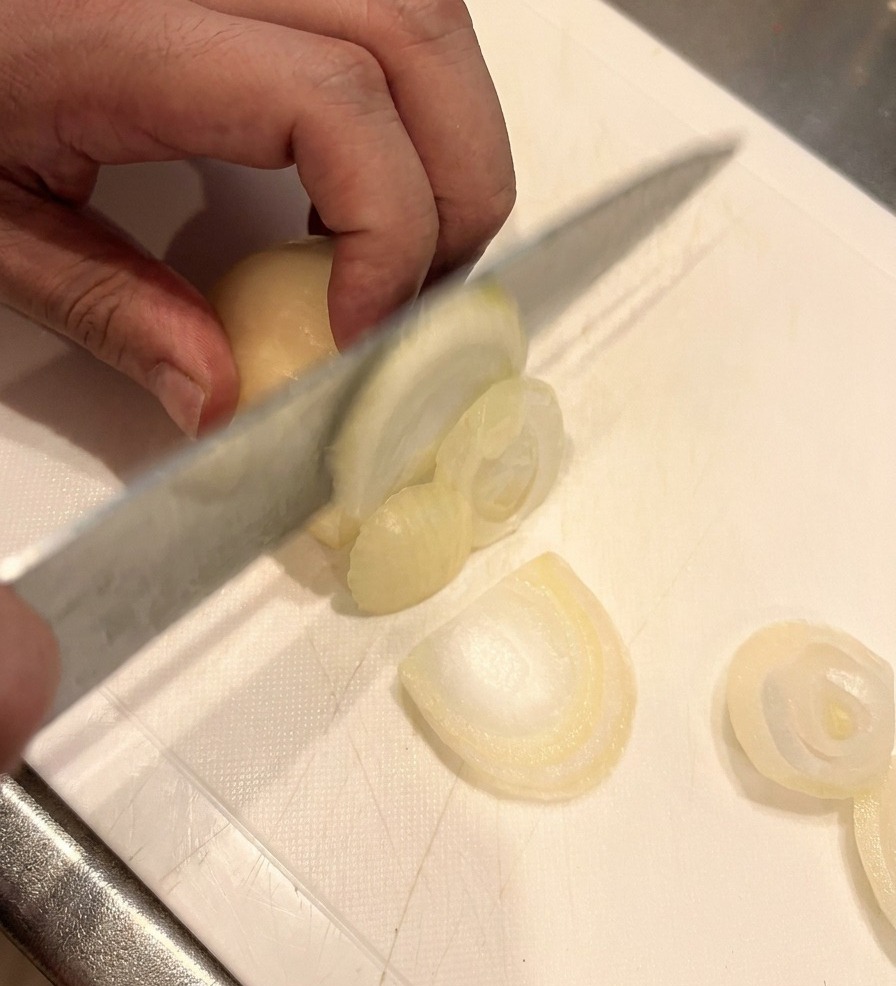Un coltello affilato taglia una cipolla bianca a fettine sottili su un tagliere bianco, con una mano che tiene saldamente la cipolla.