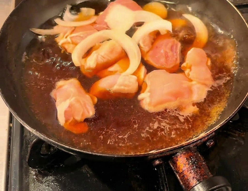 Pezzi di pollo e cipolle affettate che cuociono in un brodo scuro alla salsa di soia in una padella nera.