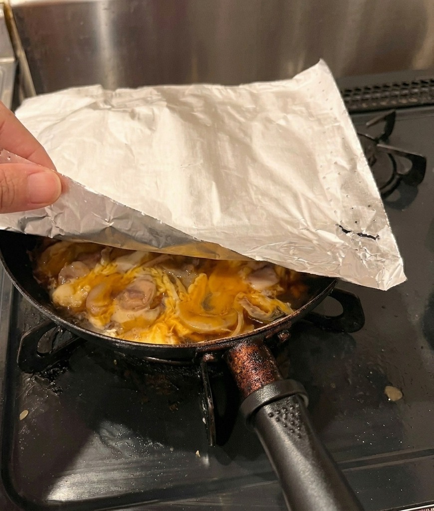 Una mano che posiziona un foglio di carta forno sopra una padella con pollo e uova parzialmente cotte.