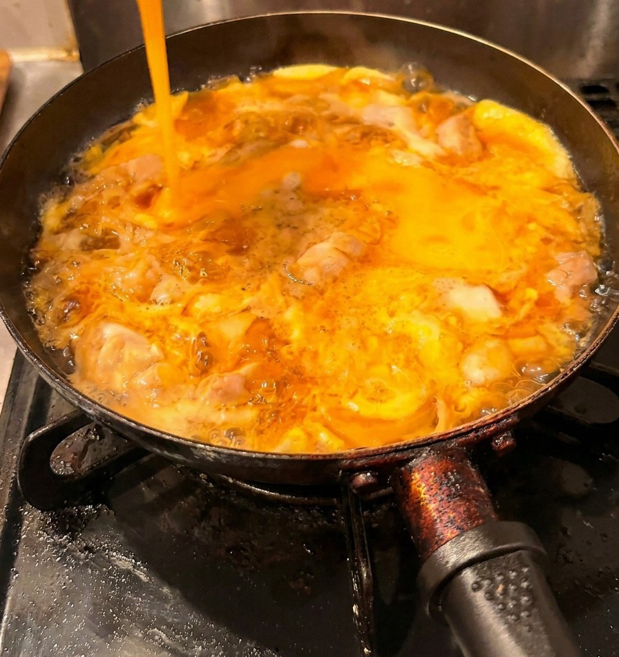 Un filo di uova sbattute giallo brillante versato in padella sopra il pollo e il primo strato di uovo già cotto.
