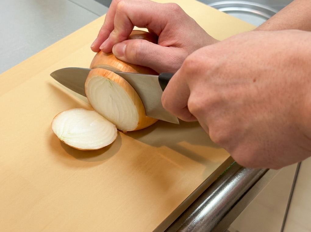 Una persona che taglia a metà longitudinalmente una cipolla gialla intera e sbucciata su un tagliere chiaro.