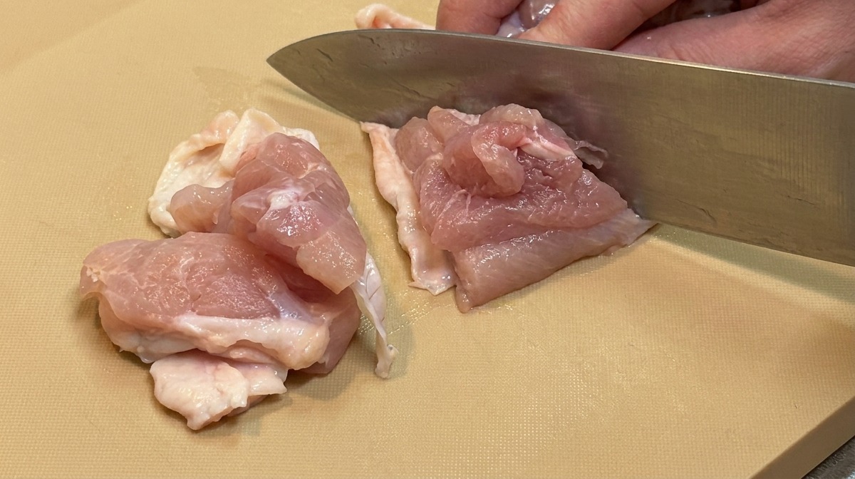 Pollo crudo disossato tagliato a cubetti uniformi della grandezza di un boccone con un grande coltello da cucina.