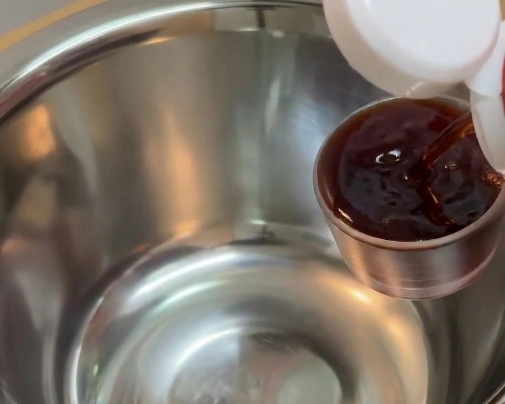 Salsa di soia scura versata da un piccolo misurino di metallo in una ciotola di acciaio inossidabile più grande.
