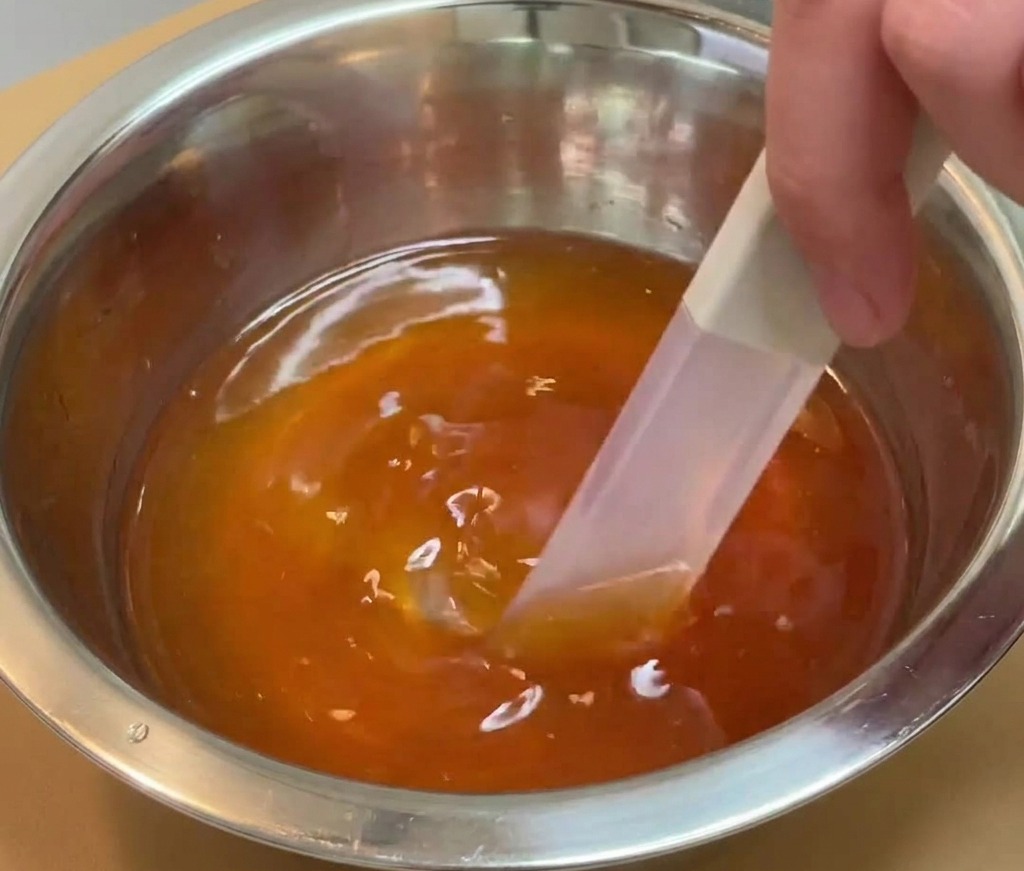 Una mano che usa una spatola per mescolare un liquido di condimento scuro e traslucido in una ciotola di acciaio inossidabile.