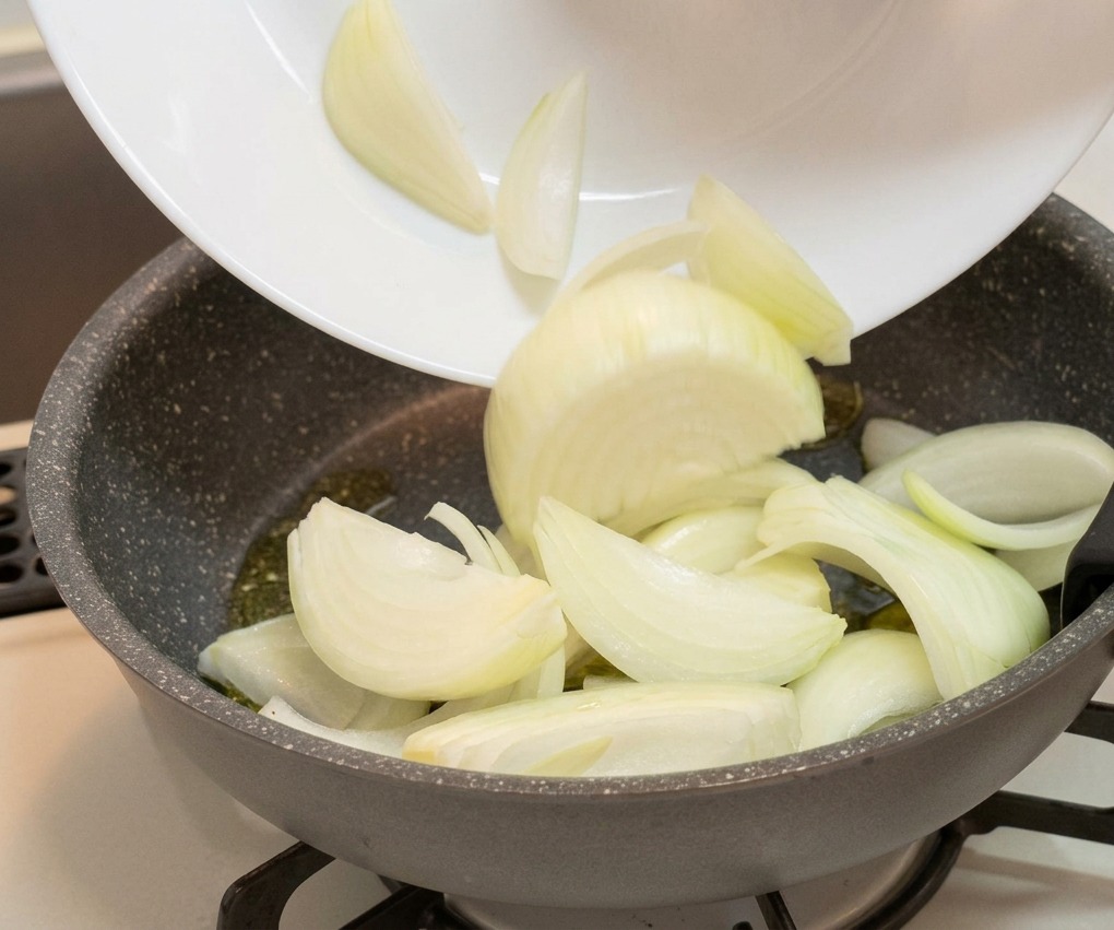Spicchi di cipolla bianca trasferiti da un piatto bianco a una padella antiaderente grigia.