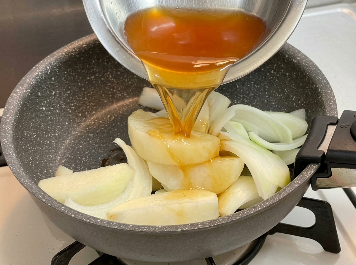 Liquido di condimento scuro versato da una ciotola di metallo sopra un letto di spicchi di cipolla in una padella.