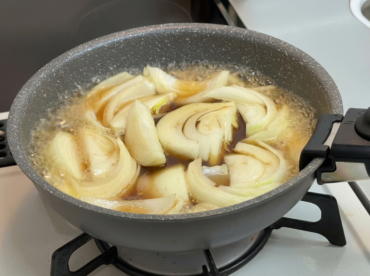 Cipolle che sobbollono in un brodo marrone scuro e spumeggiante allinterno di una padella grigia su un piano cottura.