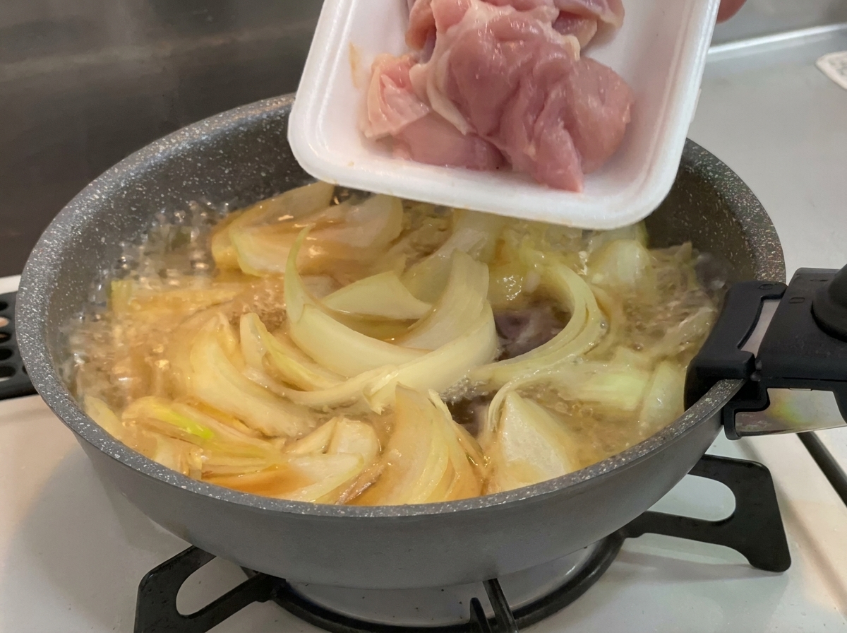 Pezzi di sovracoscia di pollo crudi e rosa aggiunti da un vassoio di polistirolo bianco in una padella con cipolle e brodo che sobbollono.