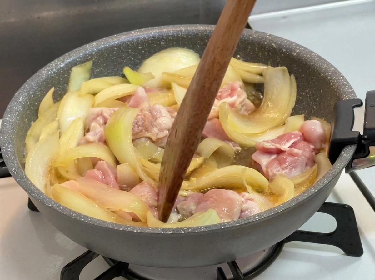 Una spatola di legno che mescola pezzi di pollo parzialmente cotti e cipolle in un liquido scuro che sobbolle in padella.