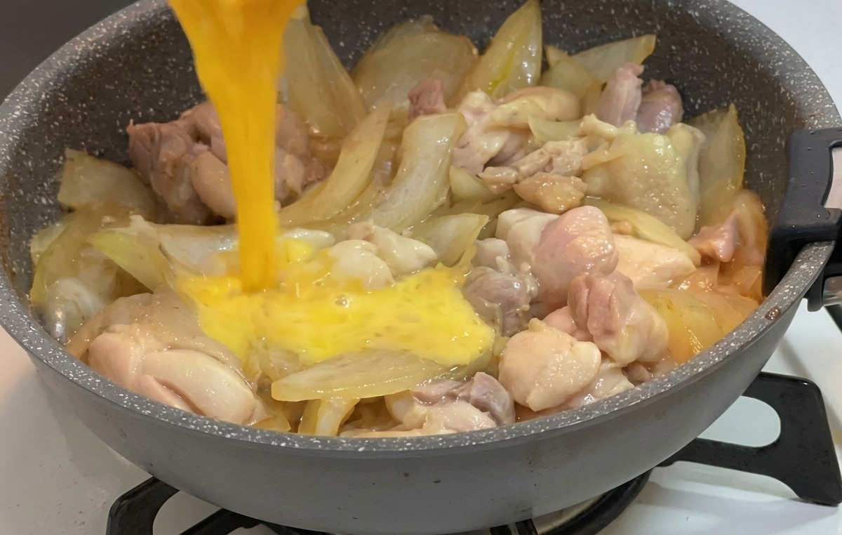 Uovo sbattuto versato da una piccola ciotola su pollo e cipolle che sobbollono in una salsa.