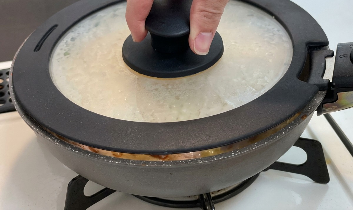 Una mano che posiziona un coperchio di vetro con un pomolo nero su una padella contenente pollo, cipolle e uova.