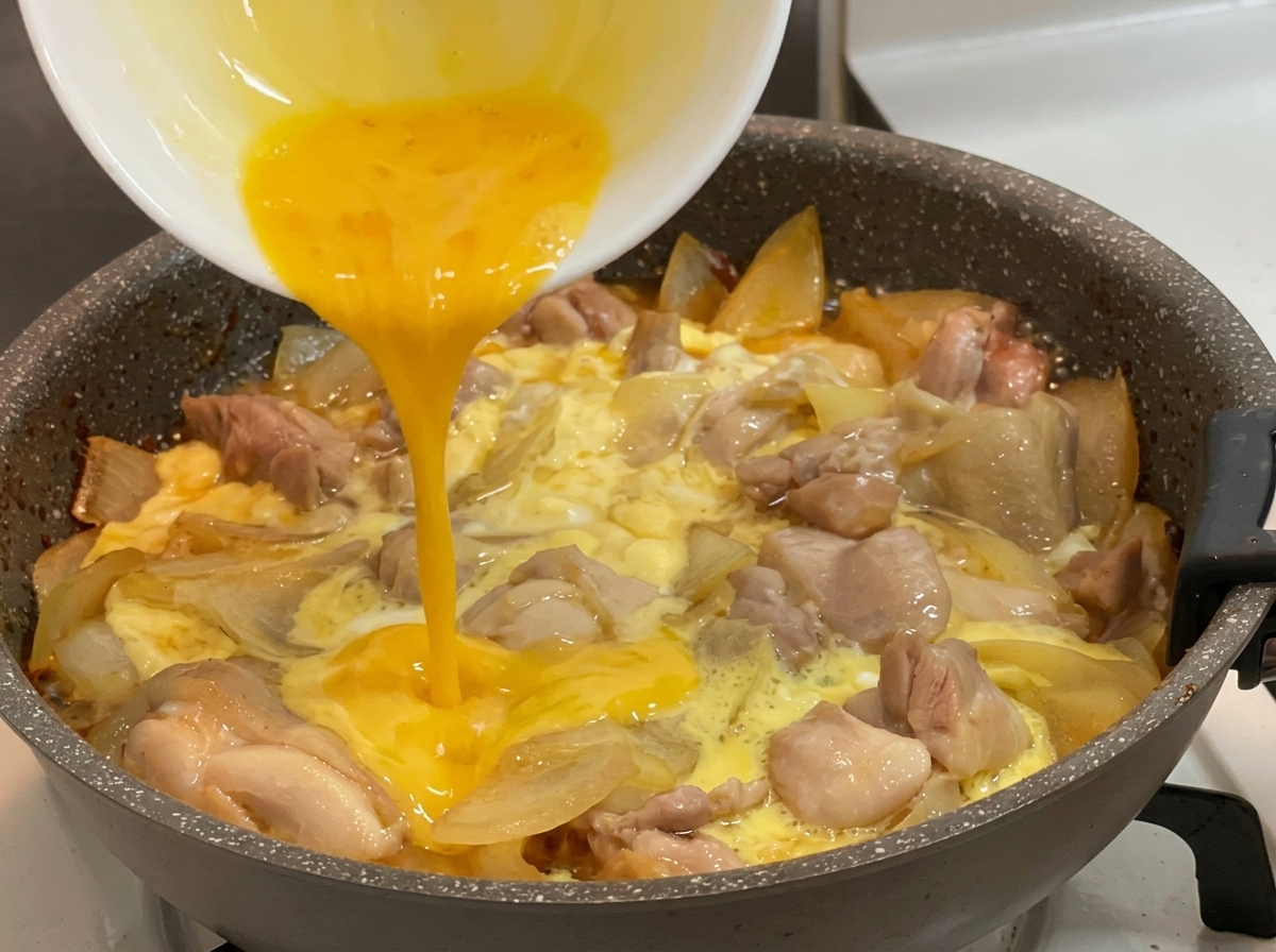 Uova sbattute di un giallo acceso versate da una ciotola bianca in una padella calda piena di pezzi di pollo, cipolle affettate e uova parzialmente rapprese.