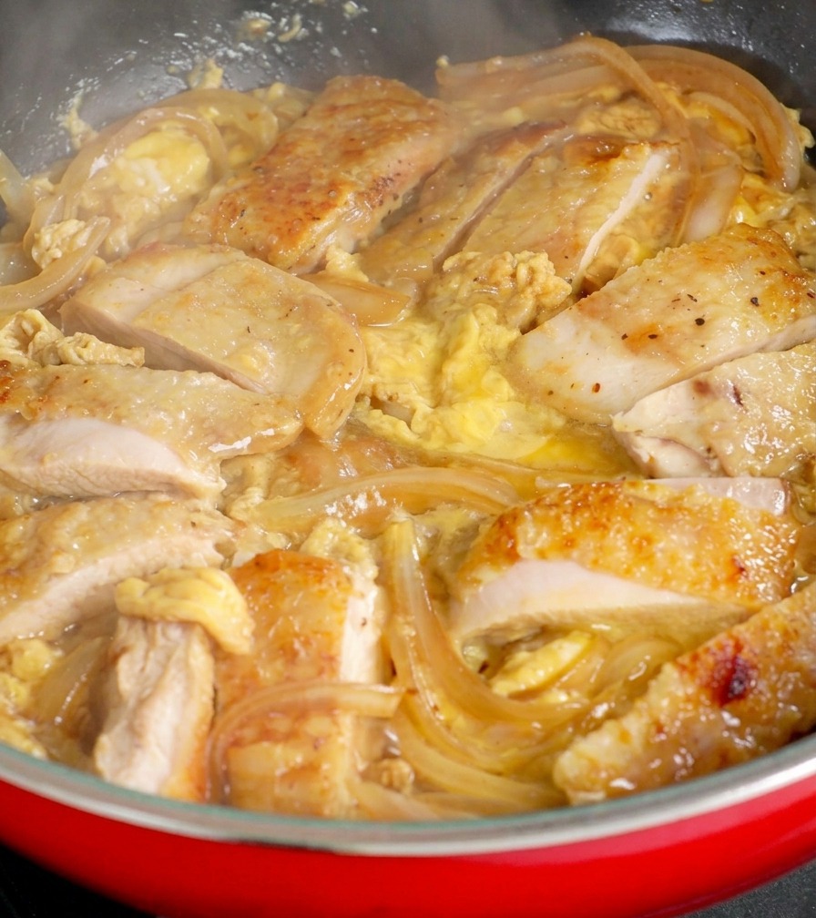 Pezzi di pollo dorato tra cipolle traslucide in una padella rossa, con uovo crudo versato sopra per creare una consistenza cremosa.