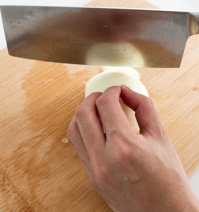 Una mano che usa un grande coltello in acciaio inox per affettare una cipolla bianca a strisce sottili su un tagliere di bambù.