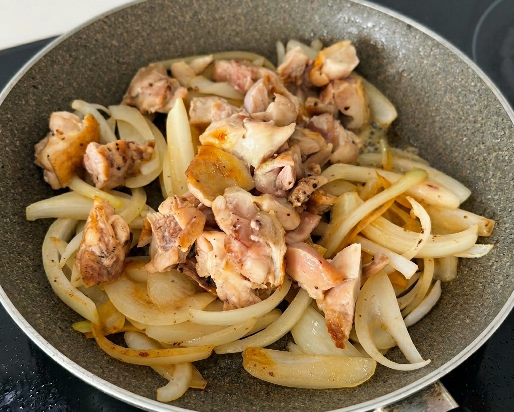 Fette di cipolla saltate mescolate con pezzi di pollo dorati e cipollotti in una padella antiaderente.