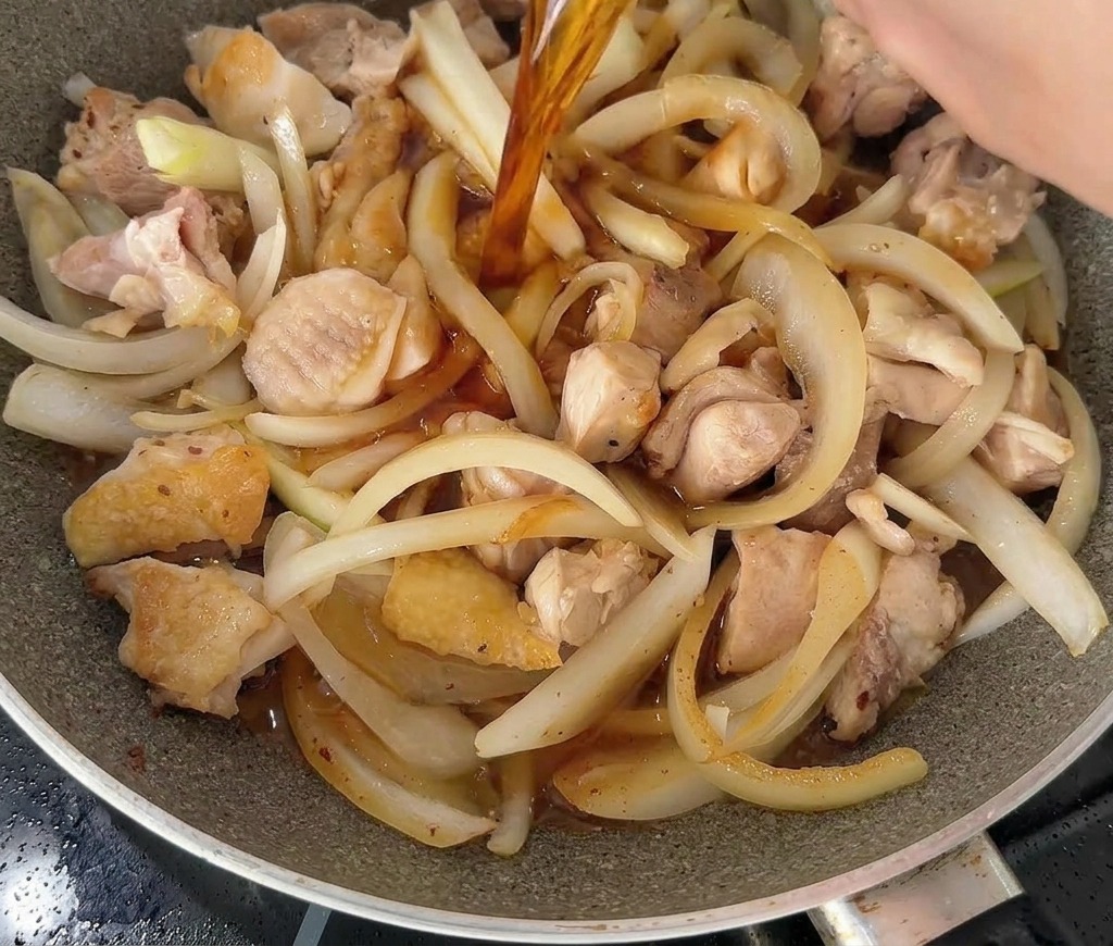 Un liquido scuro e saporito versato da una ciotola bianca in una padella con pezzi di pollo e cipolle.