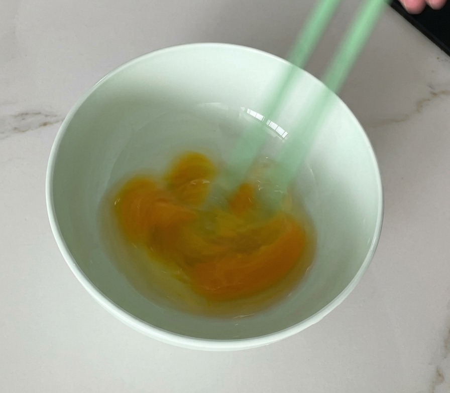 Un paio di bacchette verdi che sbattono uova crude in una ciotola verde chiaro, mostrando una miscela marmorizzata di tuorlo e albume.