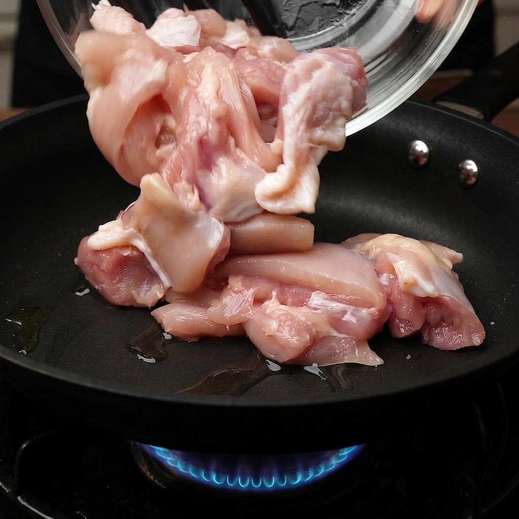 Pezzi di pollo crudo versati da una ciotola di vetro in una padella calda con una fiamma a gas blu visibile sotto.