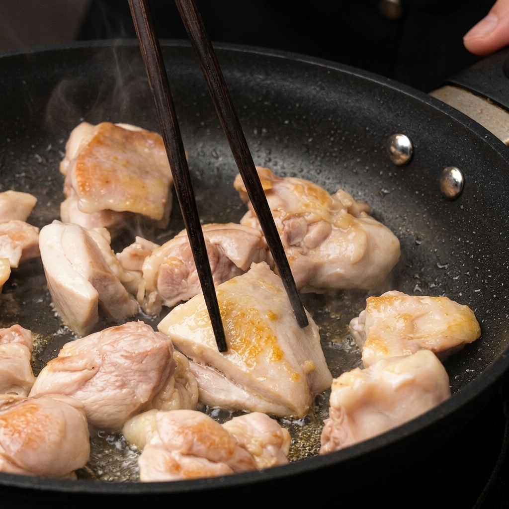 Pezzi di pollo che si dorano in padella, girati con bacchette di legno scuro.