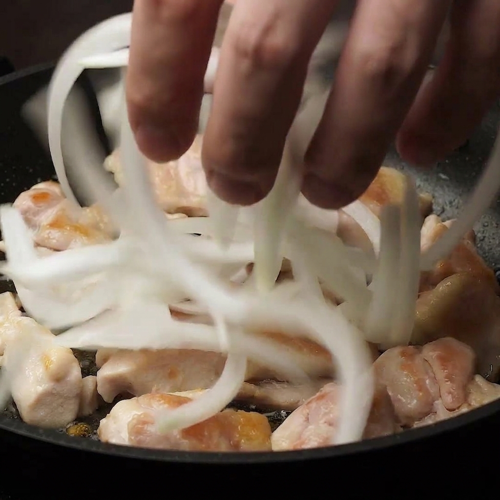 Una mano che aggiunge cipolle bianche affettate sottilmente in una padella sopra pezzi di pollo parzialmente cotti.