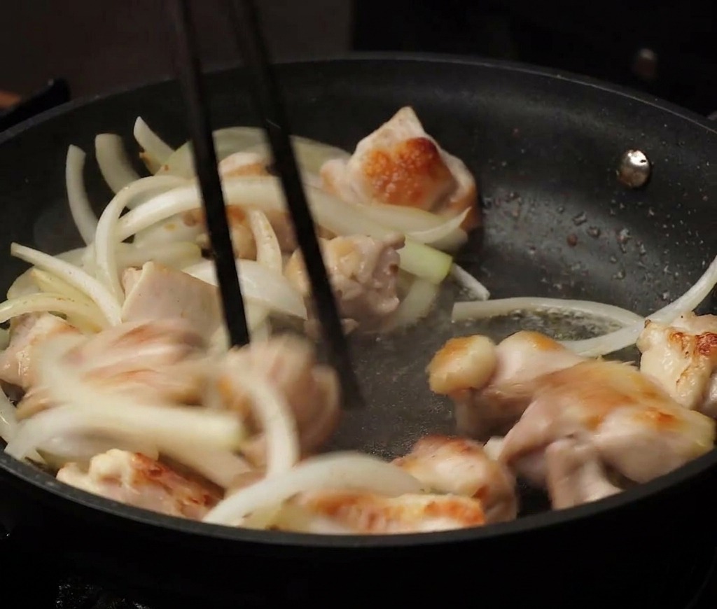 Bocconcini di pollo e cipolle affettate saltati insieme in una padella antiaderente nera usando le bacchette.
