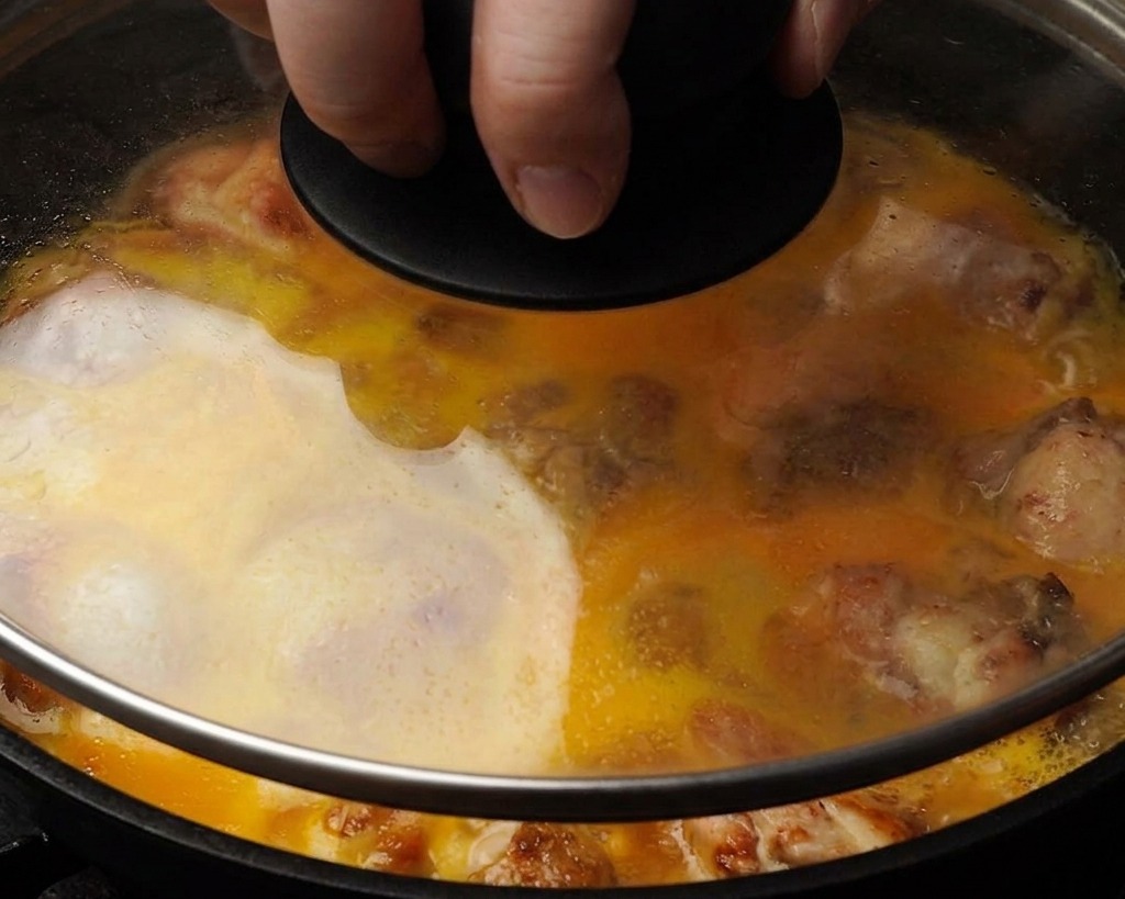 Primo piano di una padella con coperchio di vetro che mostra luovo liquido giallo che copre parzialmente i pezzi di pollo mentre cuociono a vapore.