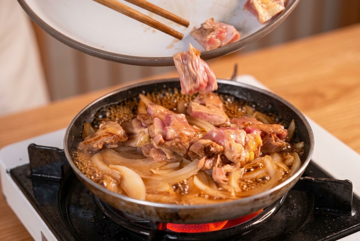 Pezzi di pollo scottato aggiunti con le bacchette in una padella con cipolle e brodo dashi in ebollizione.