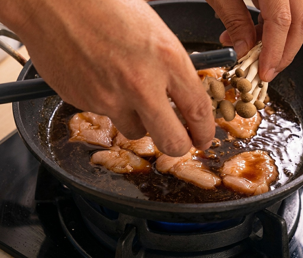 Mani che adagiano un gruppo di funghi shimeji in una padella con pollo e salsa scura in ebollizione.